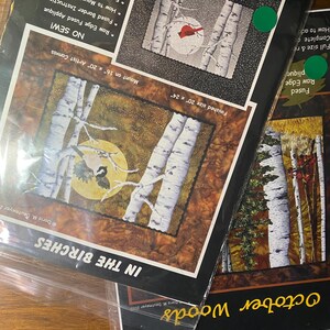 May include: Several packaged craft kits are displayed, featuring nature-themed artwork. The kits include patterns for projects like "In the Birches" and "October Woods." The packaging highlights features such as fused applique and artist canvas mounting.