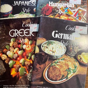 May include: A stack of vintage cookbooks featuring international cuisine. Titles include "Japanese Style," "Hungarian Style," "Greek Style," and "German Style." Each cover displays a colorful image of a dish, with text and price tags visible.