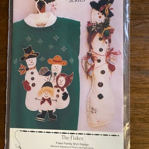 Może przedstawiać: Opakowanie z zielonym wzorem bluzy i pluszowym bałwanem. Wzór bluzy przedstawia rodzinę bałwanów w kapeluszach. Tekst brzmi "The Flakes" i "25" 1-Piece Stuffed Snowman with Old Quilt Mittens and Fur Hat."