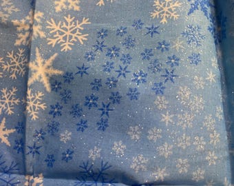 May include: Close-up of a light blue fabric with a repeating snowflake pattern. The snowflakes are in shades of white and blue, with some areas featuring glitter accents. The fabric appears to be folded, creating shadows and texture.