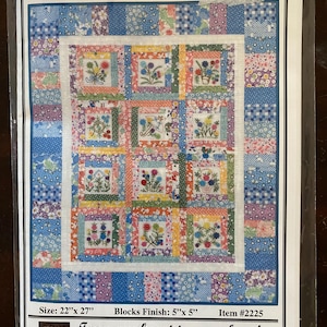 Pode incluir: Um padrão de colcha intitulado "Abby Lynn's Button Bouquet" com um design floral. A colcha apresenta um patchwork de quadrados com motivos florais bordados em várias cores. As dimensões são 56 cm x 69 cm com blocos de 13 cm x 13 cm. O texto "From my heart to your hands" também é visível.