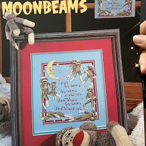以下が含まれることがあります： 「I see the moon, the moon sees me. God bless the moon, God bless me.」というテキストと、猿のグループが描かれた額装されたクロスステッチパターン。パターンのタイトルは「Monkeys & Moonbeams」で、Donna Vermillion Giampaの作品です。
