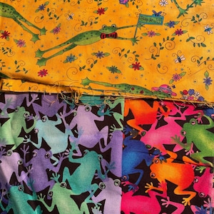 May include: Three fabric swatches with a frog theme. The top swatch is yellow with green frogs, flowers, and the text "Keep on Joy". The middle swatch is purple with green frogs on a black background. The bottom swatch is pink with orange frogs on a black background.