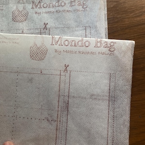 May include: Two sheets of light-colored fabric with the text "Mondo Bag" and "By Mattie Rhoades Haines" printed in a dark red color. The fabric has a dotted texture and a bag illustration. Dotted lines and scissor icons are also visible.