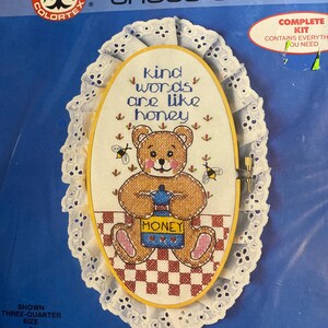May include: A cross-stitch kit featuring a teddy bear holding a honey pot, with the text "Kind words are like honey." The design is framed in a wooden hoop, surrounded by white lace. Finished size is 7.5" x 11".
