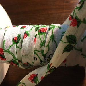 May include: A white fabric ribbon with a repeating pattern of red strawberries and green leaves. The ribbon is rolled up and tied in a knot.