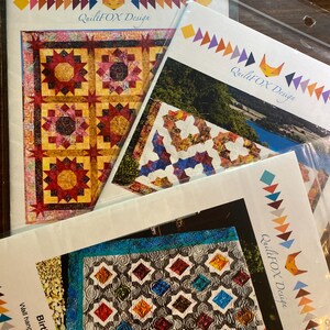 May include: Three QuiltFOX Design quilt patterns in clear plastic sleeves. The top left shows a yellow and red star quilt. The top right shows a multi-colored geometric quilt. The bottom shows a Birthstones quilt, 60 inches by 92 inches.