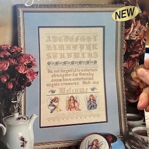 Op de afbeelding: Een ingelijste kruissteeksampler met de titel "Angels Unawares" met een alfabet, een bijbelvers en engelenillustraties. De sampler zit in een blauwe lijst met een gouden rand. Een kleine, ronde, ingelijste kruissteek van een engel is ook zichtbaar.
