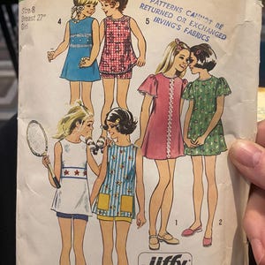 May include: Vintage Simplicity sewing pattern 5597 for girls' clothing, featuring illustrations of various outfits. The pattern includes a blue dress, red gingham shorts set, pink dress, green floral dress, and more. Size 8, breast 27 inches.