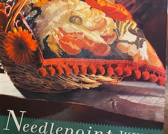 May include: A book titled "Needlepoint" by Karen Elder. The book cover features a close-up of a needlepoint pillow in a wicker basket. The pillow has a floral design with red, yellow, and white flowers. The book cover also includes the text "The Potter Needlework Library" and "Techniques, Projects, Patterns, Motifs."