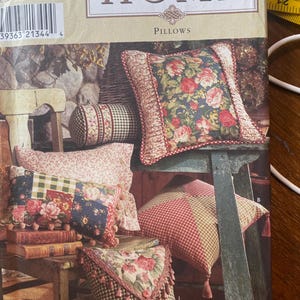 May include: A Simplicity Home sewing pattern envelope for pillows featuring various designs including floral prints, gingham checks, and patchwork patterns with decorative trim, tassels, and pom-poms, with a yellow measuring tape visible at the top.