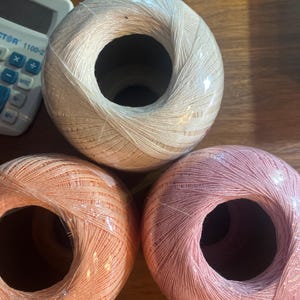 Peut inclure: Trois pelotes de fil rondes enveloppées dans un film plastique transparent. La pelote du haut est de couleur crème, celle en bas à gauche est pêche clair et celle en bas à droite est rose tendre. Une calculatrice blanche est visible en haut.