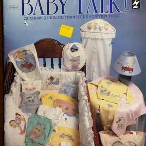 Puede incluir: Una portada de libro con el título "Baby Talk!" y el subtítulo "20 transferencias de planchado increíbles para pequeños". La portada presenta una cuna con varios artículos para bebés, incluyendo un babero, una manta, una lámpara y una bolsa de mano. Los artículos están decorados con diseños lindos con temática de bebés.