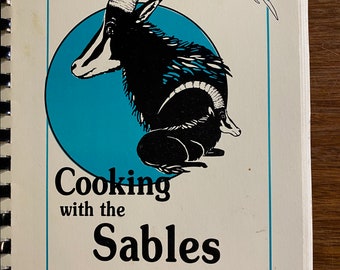 May include: A cookbook cover featuring a black and white illustration of a sable antelope against a blue circular background. The text on the cover reads "Cooking with the Sables" and "A Sables Project to Benefit the Educational Programs of Safari Club International."