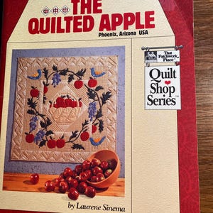 May include: A book titled "The Quilted Apple" with a quilt design featuring a basket of apples, vines, and birds. The book is part of the "Quilt Shop Series" and is by Laurene Sinema.