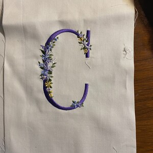 May include: A white fabric with a purple embroidered letter C surrounded by blue and yellow flowers.