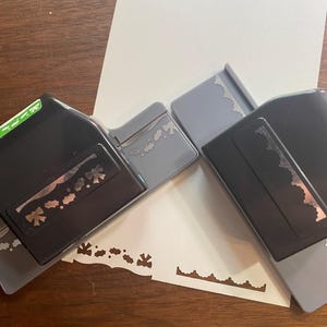 Puede incluir: Dos perforadoras de papel negras y grises con diseños de borde decorativos, junto a una hoja de papel blanca con patrones perforados. Las perforadoras tienen diferentes patrones decorativos, incluyendo lazos y bordes festoneados. El fondo es una superficie de madera.