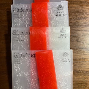 May include: Stack of Cricut Cuttlebug embossing folders with red plastic shims. The folders are clear with floral patterns and the Anna Griffin logo. The text on the folders includes "Modern Vines", "Blossom Dance", "Mayfair Floral", and "Treillage".