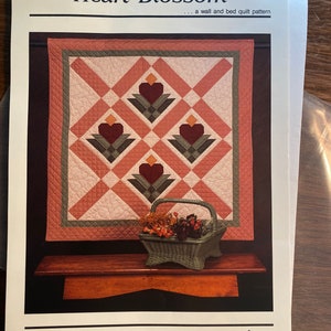 May include: A quilt pattern named "Heart Blossom" by thimbleberries, designed for walls and beds. The quilt design showcases a geometric pattern with heart-shaped floral motifs in red, green, and peach. A wicker basket with dried flowers is positioned below the quilt.