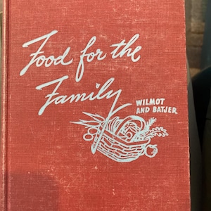 以下が含まれることがあります： 「Food for the Family」と書かれた赤い表紙のヴィンテージ料理本。タイトルは薄い青色の筆記体で、食べ物のバスケットのイラストが描かれています。著者の名前、Wilmot and Batjerも表示されています。