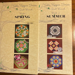 Op de afbeelding: Twee open boekjes getiteld "Table Topper Series" van "The Quilt Branch". Boek 1 is gelabeld "Spring" en Boek 2 is gelabeld "Summer". Elk boek bevat kleurrijke quiltontwerpen, waaronder bloemen- en geometrische patronen.