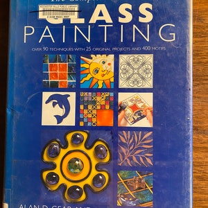 May include: The Complete Guide to Glass Painting book cover features a blue background with colourful glass painting projects. The book title is in white text with a yellow sun, a dolphin, a floral design, and a mosaic tile design.