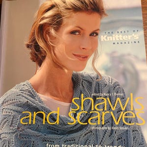 May include: The Best of Knitter's Magazine book cover featuring a woman wearing a blue knitted shawl with a lace pattern. The title of the book is "Shawls and Scarves: From traditional to trendy... with easy to follow instructions."