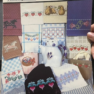 May include: A collection of tea towel embroidery patterns featuring various designs, including a unicorn, a cat, pears, strawberries, and hearts. The patterns are from Hickory Hollow Tea Towel Treasury, a cross-stitch pattern book.