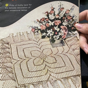 以下が含まれることがあります： 「Ruffled Doilies」というタイトルのヴィンテージ本の表紙。かぎ針編みのドイリーと花の配置が特徴です。表紙のテキストには「Frills of frothy lace for the delicate decoration of your occasional tables」と書かれています。ドイリーはスカラップエッジです。