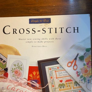 May include: A book titled "Simple-to-Sew Cross-Stitch" with a white background and a blue title. The cover features several cross-stitch projects, including a framed floral design, a framed house design, and a pillow with the word "Carol" embroidered on it.