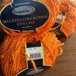 May include: Two skeins of bright orange Sensations Bellezza Collection Stellina wool blend yarn. Each skein features a black label with white text reading A Jo-Ann exclusive, Sensations, Bellezza Collection, Stellina, and Wool Blend.