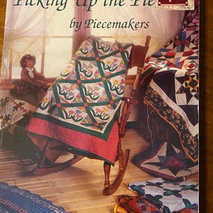 May include: A book titled "Picking Up the Pie" by Piecemakers, featuring a colorful quilt on a rocking chair, a doll, and other quilting supplies.