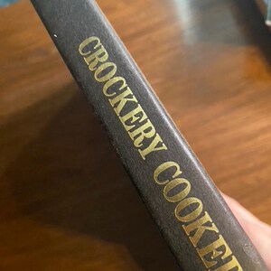 May include: A dark brown book spine with gold lettering that reads CROCKERY COOKERY. The text is oriented vertically along the spine, and a partial word is visible at the top edge of the book cover, showing the letters un's in gold font.