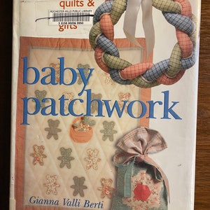 May include: Book cover with the title "baby patchwork" in large, bold letters. The cover features a braided wreath made of plaid fabric in shades of red, blue, and green, a small quilted bag with a red star, and a quilt with a pattern of teddy bears.
