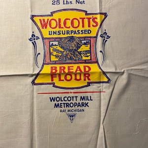 Könnte beinhalten: Ein alter weißer Mehlsack mit blauer und roter Schrift. Der Text lautet "25 Lbs. Net Wolcott's Unsurpassed Bread Flour Wolcott Mill Metropark Ray, Michigan". Der Sack zeigt eine gelbe und blaue Illustration einer Weizengarbe.