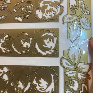 Może przedstawiać: Złote, wykrojone ozdoby kwiatowe z białymi akcentami. Obraz przedstawia kilka prostokątnych i pionowych element&oacute;w z motywami r&oacute;ż i liści. Wycięte detale tworzą efekt warstwowy, odpowiedni do scrapbookingu lub tworzenia kartek.