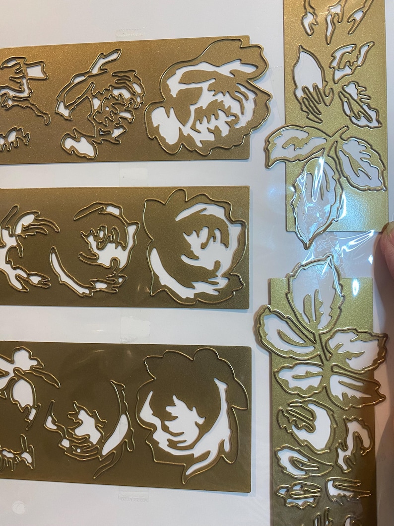 Może przedstawiać: Złote, wykrojone ozdoby kwiatowe z białymi akcentami. Obraz przedstawia kilka prostokątnych i pionowych element&oacute;w z motywami r&oacute;ż i liści. Wycięte detale tworzą efekt warstwowy, odpowiedni do scrapbookingu lub tworzenia kartek.