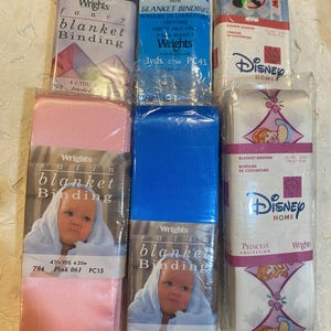 May include: Assortment of blanket binding rolls in various colors and designs. Includes pink, blue, green, and white rolls. Some rolls feature Disney characters and baby images. Each roll is packaged and labeled with the brand "Wrights".