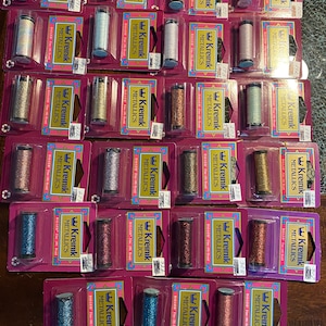 May include: A collection of 20 spools of Kreinik metallic embroidery floss in various colors, including gold, silver, copper, and blue. Each spool is labeled with the Kreinik logo and the number of the color.