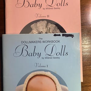 May include: Two vintage dollmaking workbooks, "Baby Dolls" by Mildred Seeley, Volume I and II. Volume II is pink, Volume I is light blue. The covers feature the title in elegant script.
