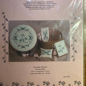 May include: A cross-stitch kit titled "Rose Garden Memories" featuring a round shaker box lid, scissors case, needlebook, and other accessories. The kit includes instructions and charts. The design includes floral and geometric patterns in green and red.