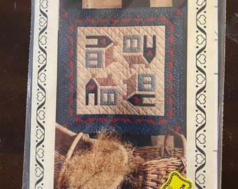 Op de afbeelding: Een pakket voor een quiltpatroon getiteld "Yankee Homestead". Het quiltontwerp heeft huis- en stermotieven in rood, blauw en bruin. Het pakket zit in een doorzichtige plastic hoes.
