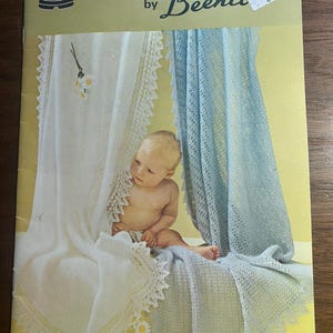 May include: Vintage book cover featuring the text "PATONS SHAWLS by Beehive." The cover displays a baby surrounded by white and light blue shawls with lace trim. The background is yellow.
