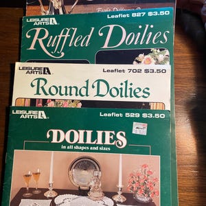 Pode incluir: Uma pilha de folhetos de padrões de crochê vintage com títulos como "Elegant Crocheted Doilies" e "Ruffled Doilies". As capas são verdes e creme, com imagens de toalhas de mesa e arranjos florais. Os folhetos são da Leisure Arts.