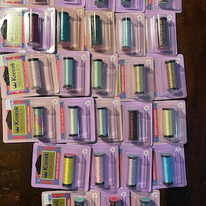 May include: A collection of 20 spools of Kreinik metallic embroidery floss in various colors, including gold, silver, green, blue, and pink. Each spool is labeled with the brand name "Kreinik" and the words "Metallic" and "Ballers".