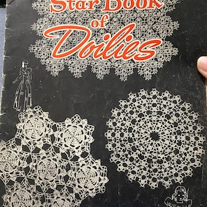 Puede incluir: Un libro vintage titulado "Star Book of Doilies" con un logotipo de estrella roja. La portada presenta un fondo negro con doilies de ganchillo blanco.