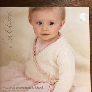 Puede incluir: Un bebé con un cárdigan de punto color crema con ribete rosa, presentado en la portada de "The little Sublime hand knit book". La portada del libro también muestra el texto "baby cashmere merino silk dk" y "15 diseños para bebés desde el nacimiento hasta los dos años."
