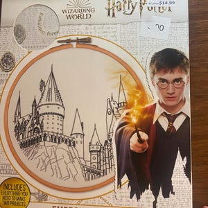 May include: A Harry Potter embroidery kit featuring a circular design of a castle and the text "EMBROIDERY KIT". The box includes an image of Harry Potter holding a wand. The kit is inspired by the Wizarding World.