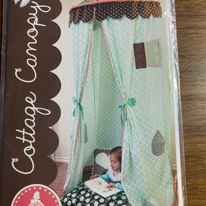 以下が含まれることがあります： ミントグリーンと白のダイヤモンド柄のコテージキャノピー。茶色とピンクのスカラップトリム付き。キャノピーには小さな窓のディテールがあり、緑のリボンで結ばれています。パッケージには「Cottage Canopy」とロゴが記載されています。