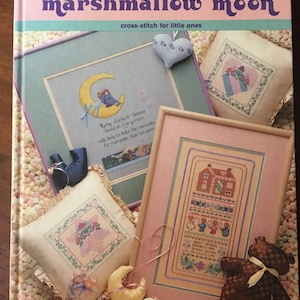 Puede incluir: Un libro titulado "Marshmallow Moon: Cross-stitch for Little Ones" con una portada rosa, azul y verde. La portada presenta varios diseños de punto de cruz, incluyendo una luna, una casa y un corazón. El libro está rodeado de pequeños malvaviscos coloridos.